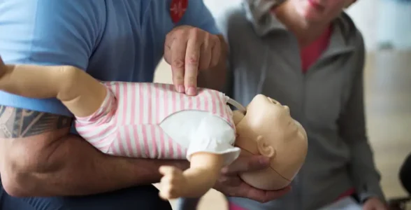 A man gives paediatric CPR to a dummy with a 4JH instructor aiding him.